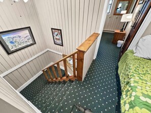 Una habitación con una escalera de madera, una cama con colcha estampada en verde y amarillo, y una mesita de noche con una lámpara.