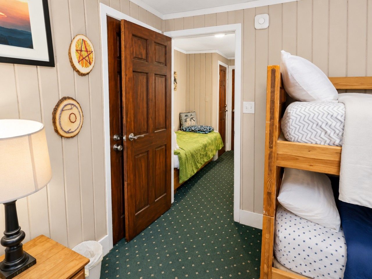 Ein Zimmer mit einem Etagenbett, einem Nachttisch aus Holz mit einer Lampe, einer Tür, die in einen anderen Raum führt, und einem gemusterten Teppich.