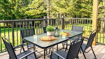 An outdoor deck with a glass table and chairs, surrounded by a wooden railing, with a potted plant and a bowl of fruit on the table.