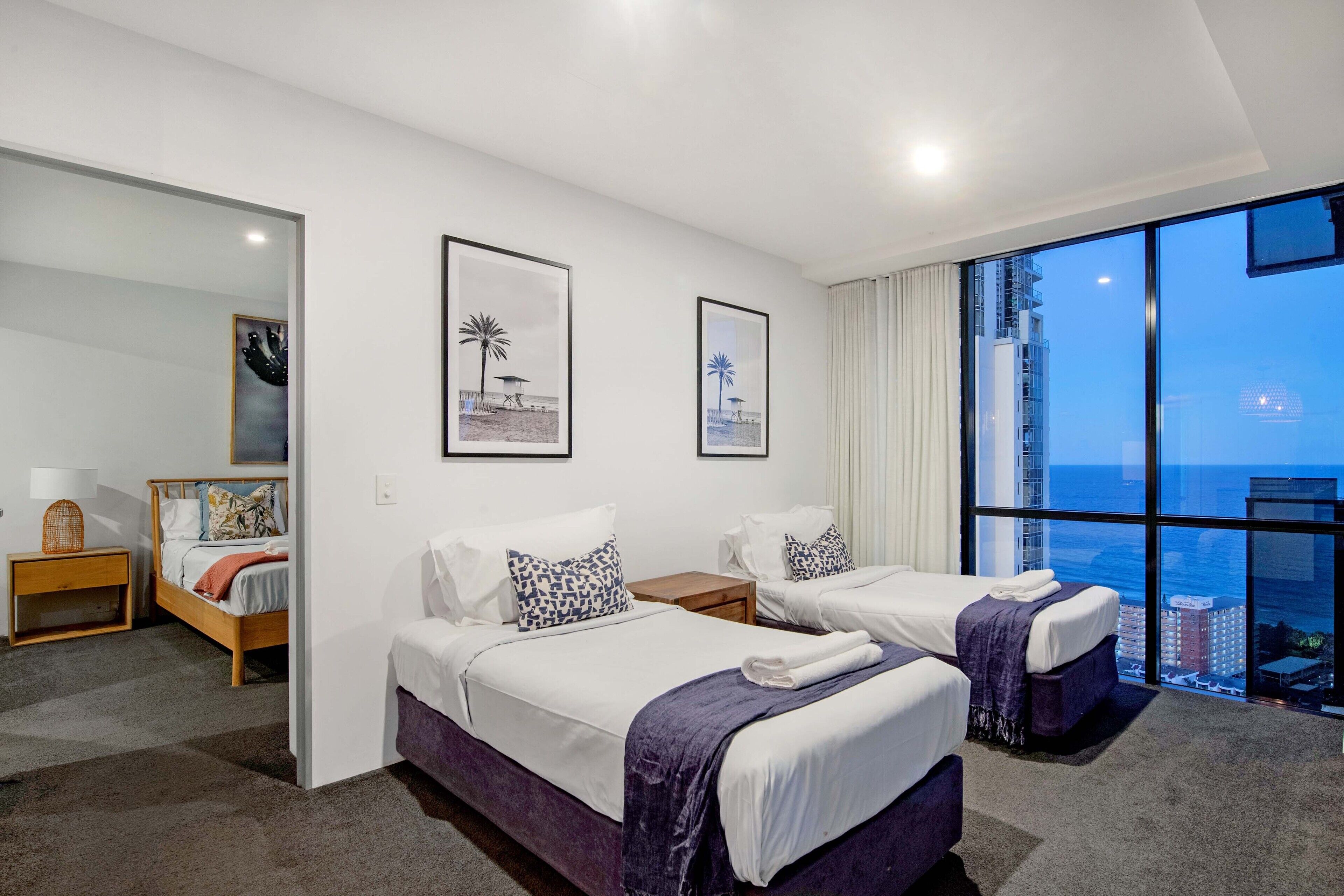 Ein Hotelzimmer mit zwei Betten, einem großen Fenster mit Stadtblick und gerahmten Bildern an der Wand.