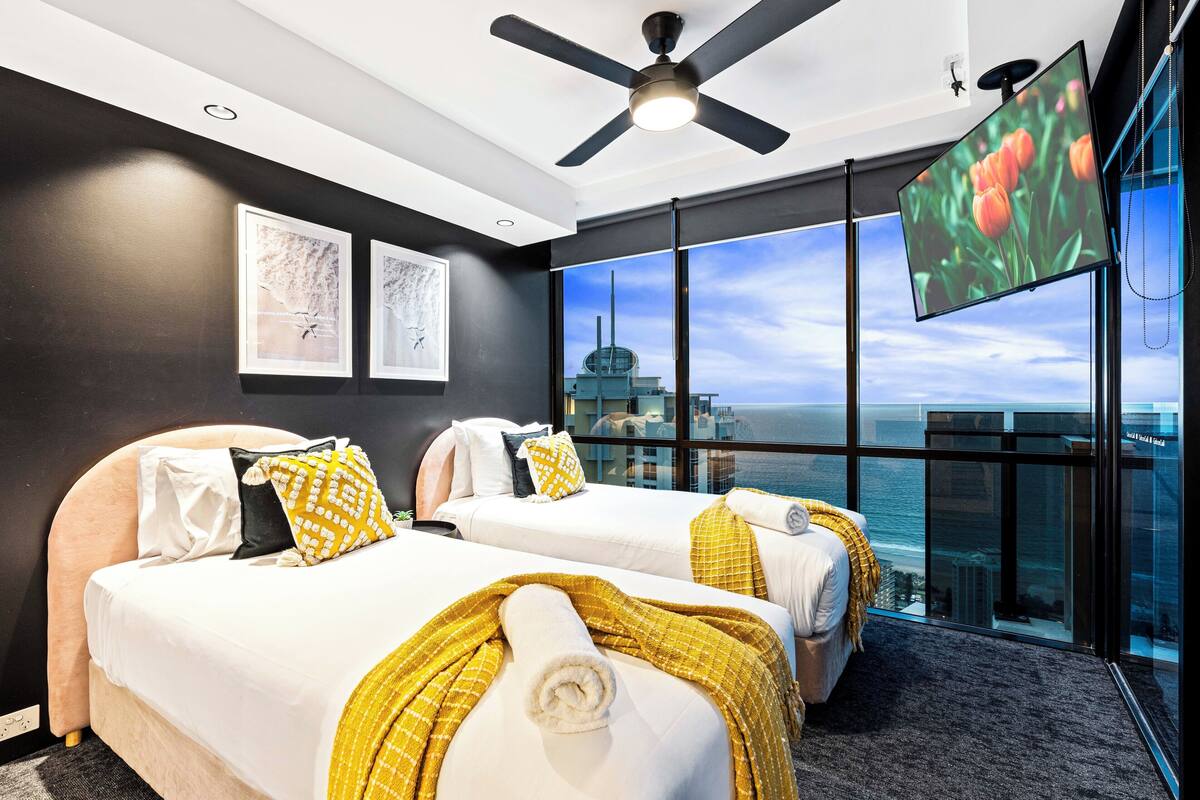 Habitación de hotel con dos camas, ventilador de techo, televisión y un amplio ventanal con vistas a la ciudad.