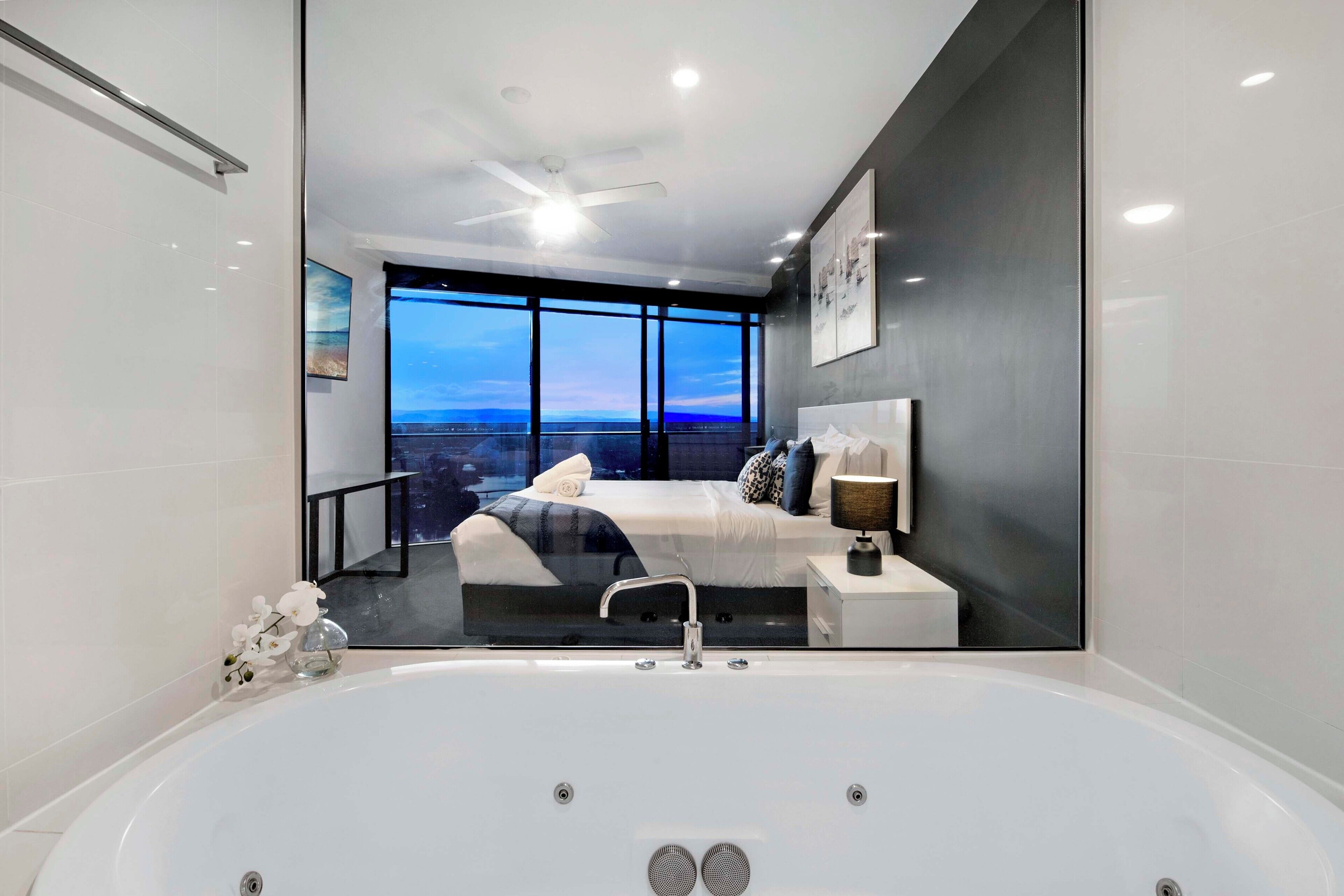 A modern bedroom with a large window, a bed, a nightstand, and a bathtub.
