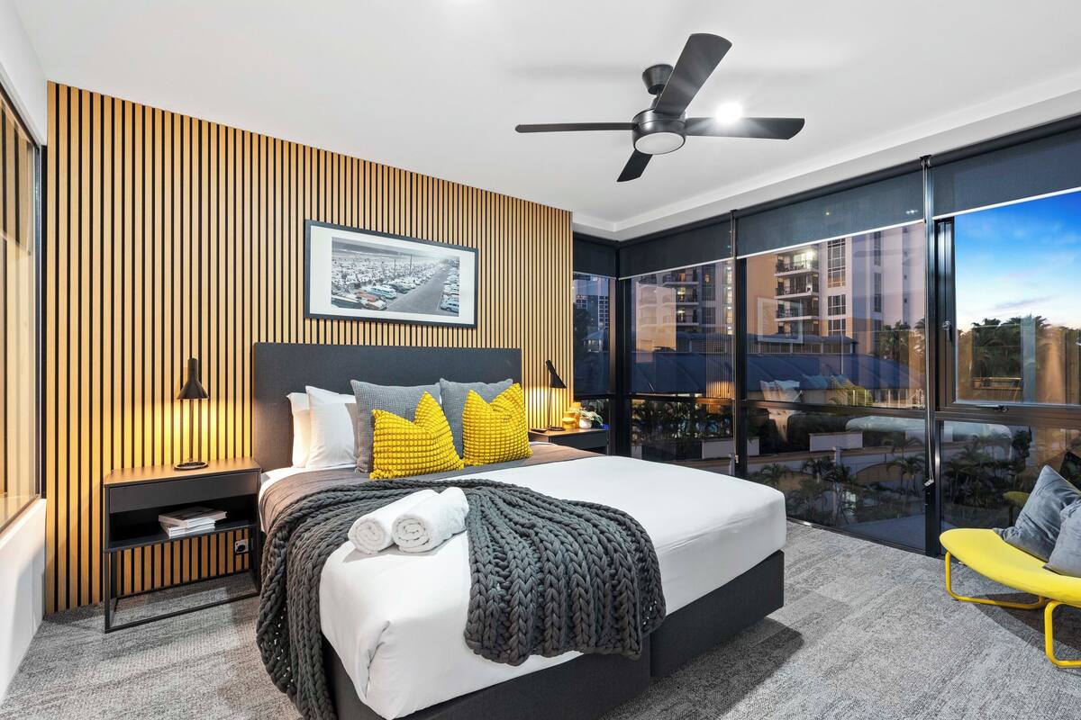 Un dormitorio moderno con una cama grande, una mesita de noche, un ventilador de techo y un balcón con vista a la ciudad.