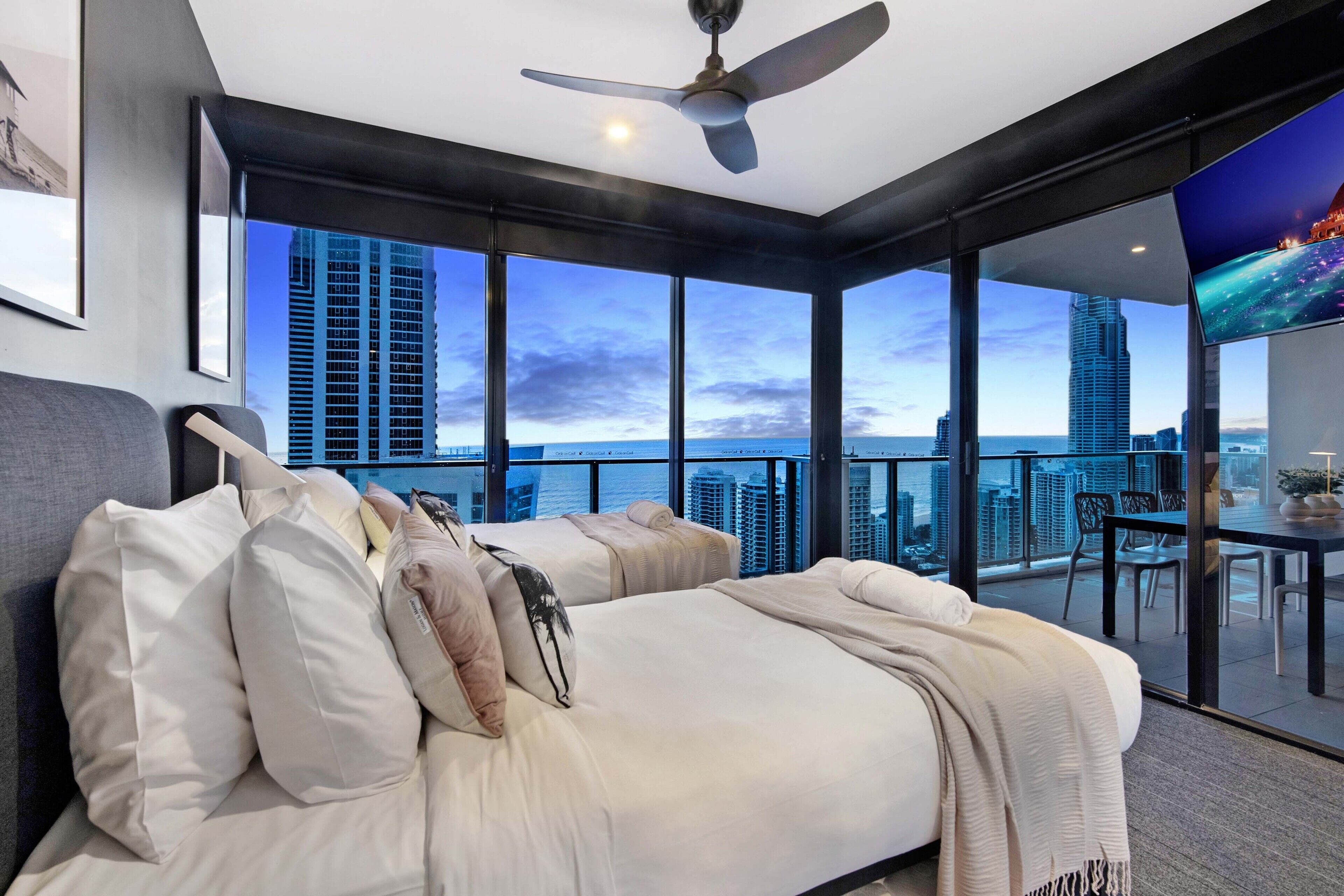 Une chambre moderne dotée d’un grand lit, d’un ventilateur de plafond et offrant une vue sur la ville grâce à de grandes fenêtres du sol au plafond.