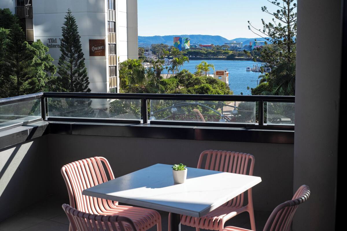 Un balcón con mesa y sillas, con vistas a un cuerpo de agua y un paisaje urbano.