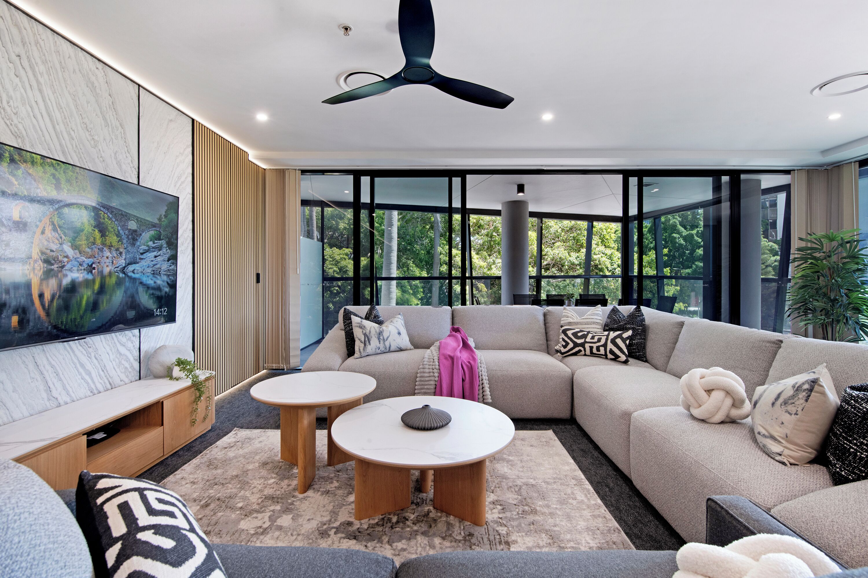 A modern living room with a sectional sofa, two round coffee tables, a flat-screen TV, and large windows offering a view of greenery.