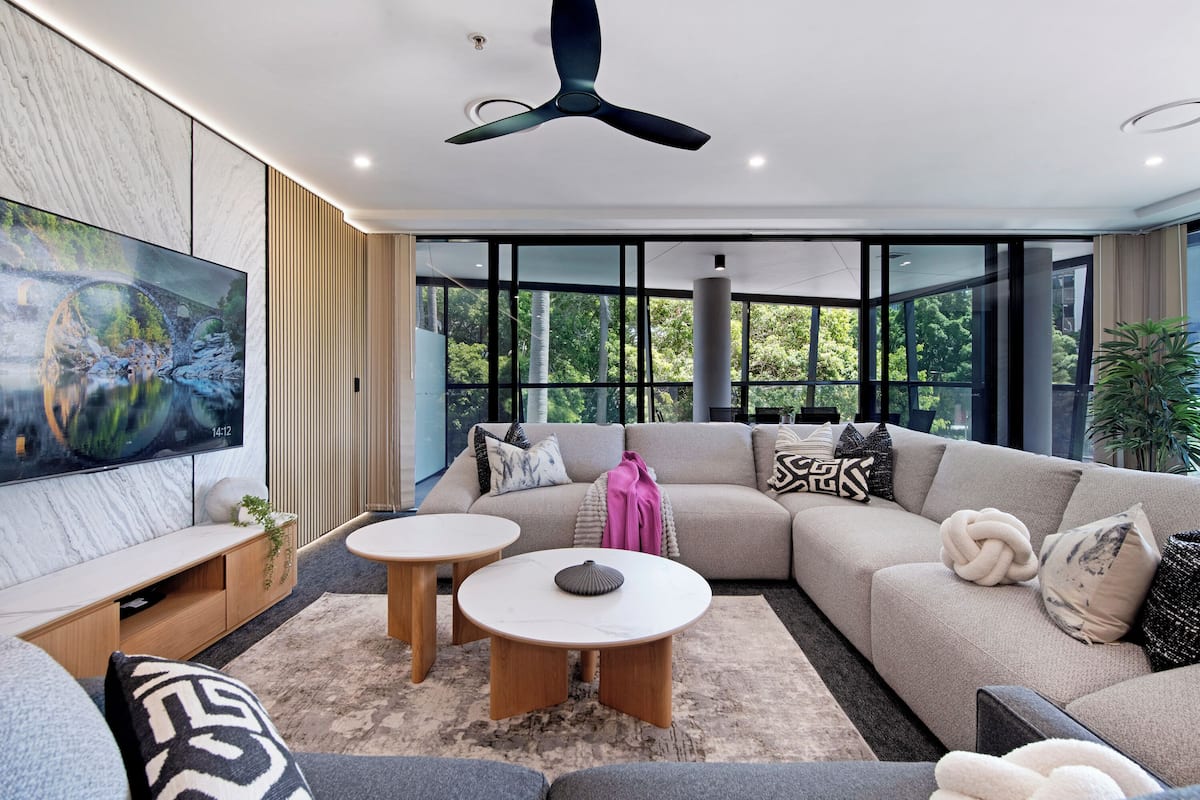 A modern living room with a sectional sofa, two round coffee tables, a flat-screen TV, and large windows offering a view of greenery.