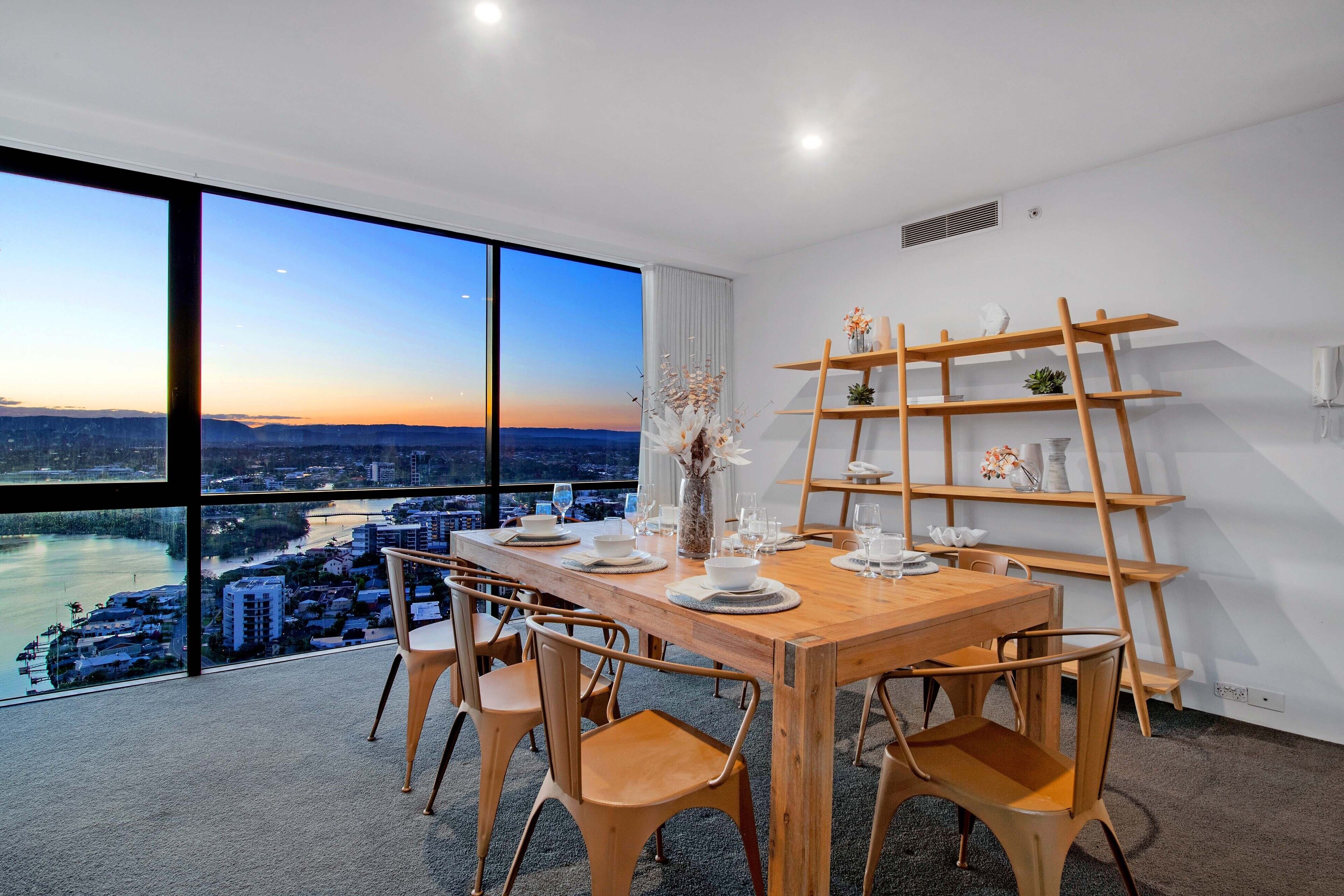 Comedor con vistas a un paisaje urbano y al río, que incluye una mesa de madera, sillas y un estante decorativo.