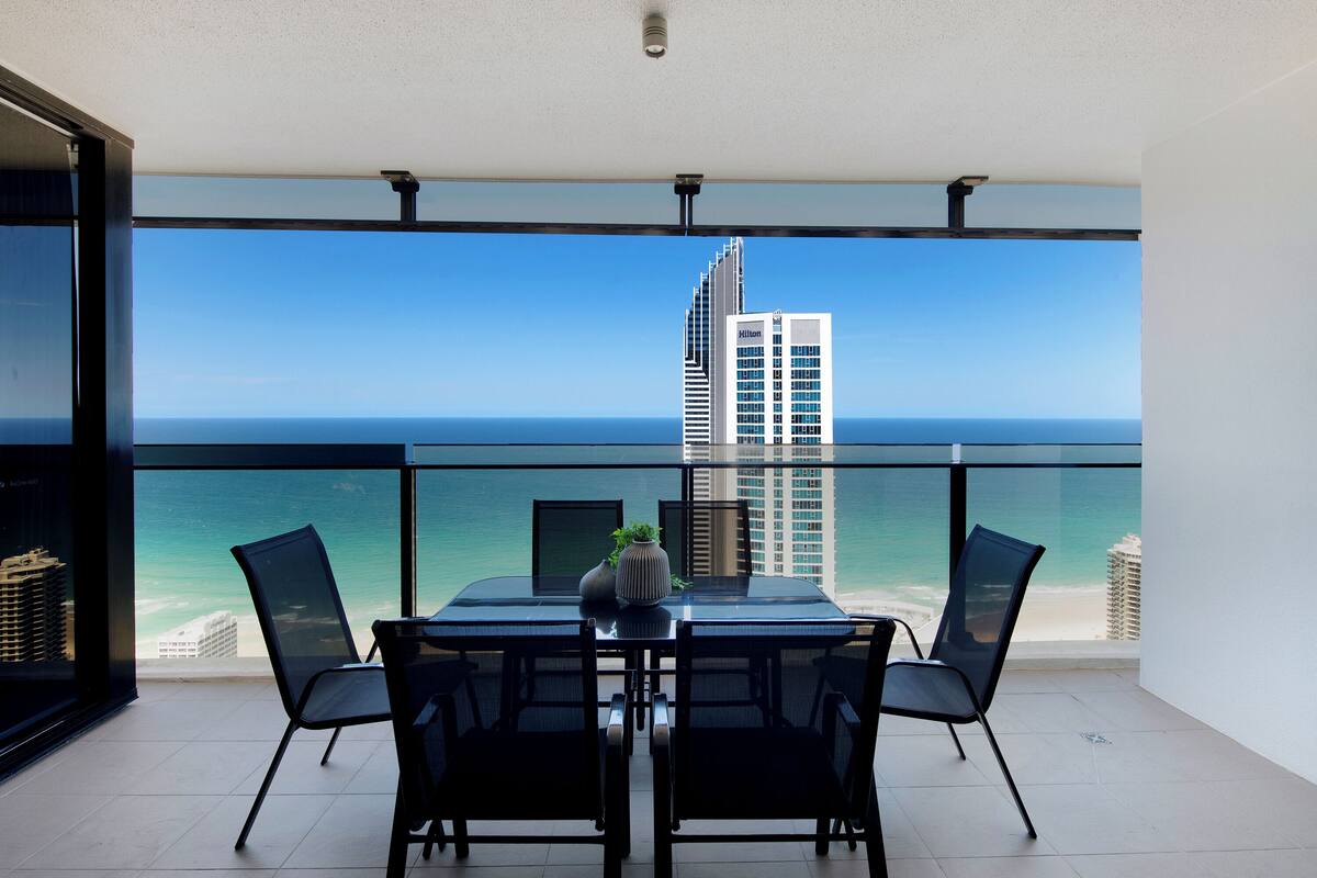 Un balcón con mesa y sillas, con vistas al océano y un edificio alto.