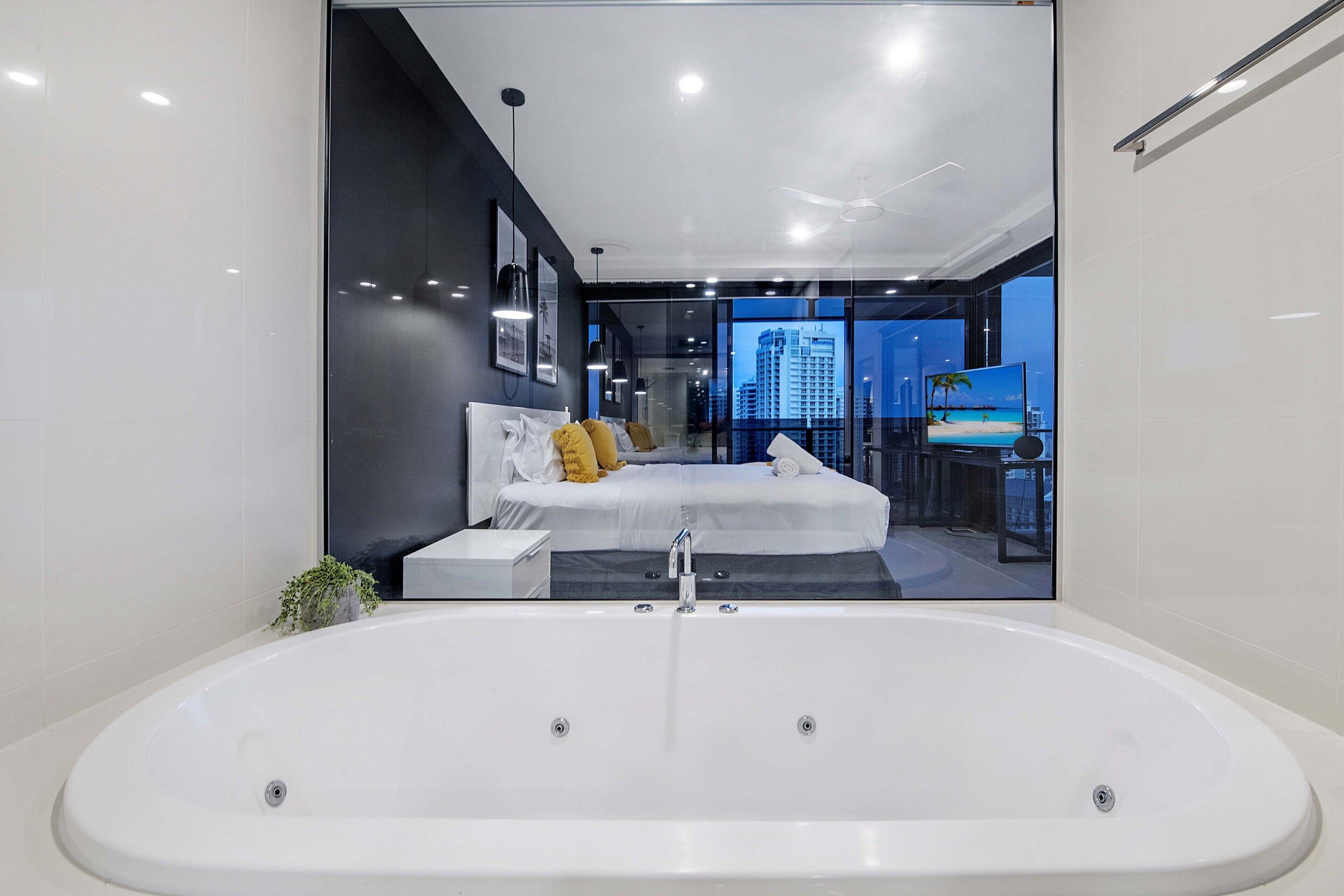 A modern bedroom with a large bathtub, a bed with white linens and yellow pillows, and a city view through large windows.