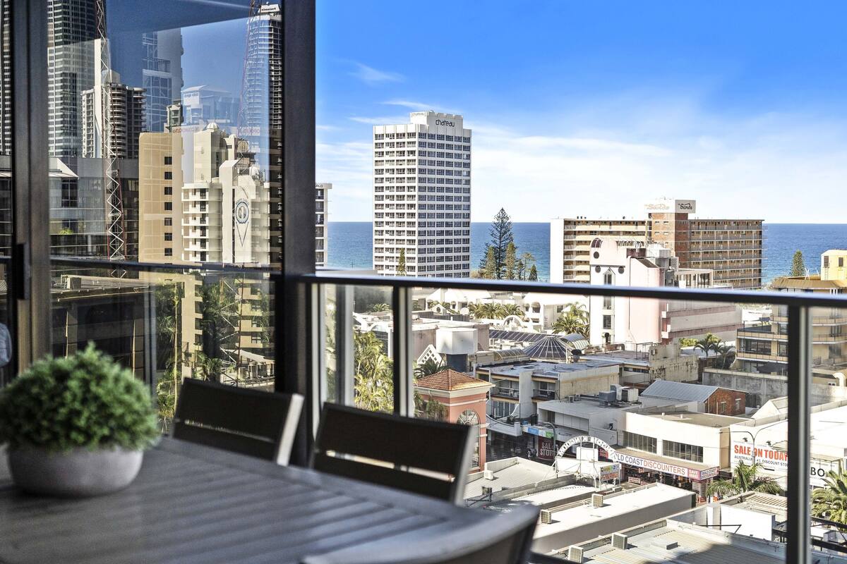 Vista panorámica de la ciudad desde un balcón con mesa y sillas, con edificios y el océano a la vista.