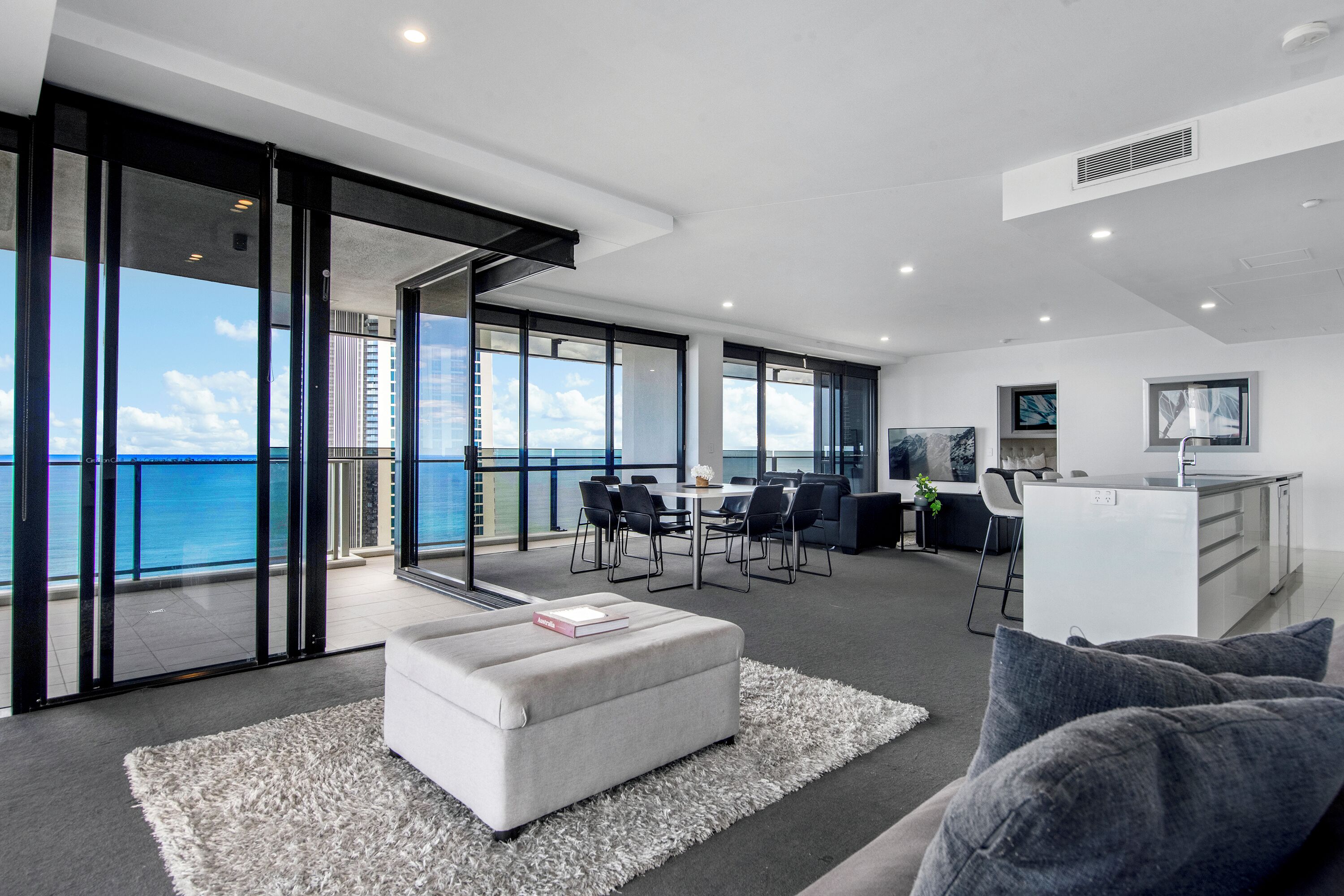 A modern living room with a large window overlooking the sea, a dining area with chairs, a kitchen with a white countertop, and a sofa with a book on it.