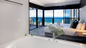Un dormitorio moderno con una bañera grande, una cama con almohadas y vistas al océano a través de amplios ventanales.