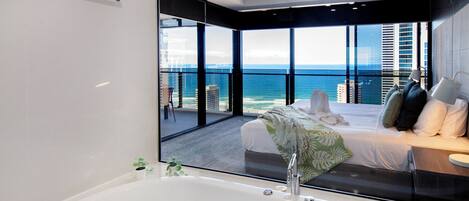 A modern bedroom with a large bathtub, a bed with pillows, and a view of the ocean through large windows.