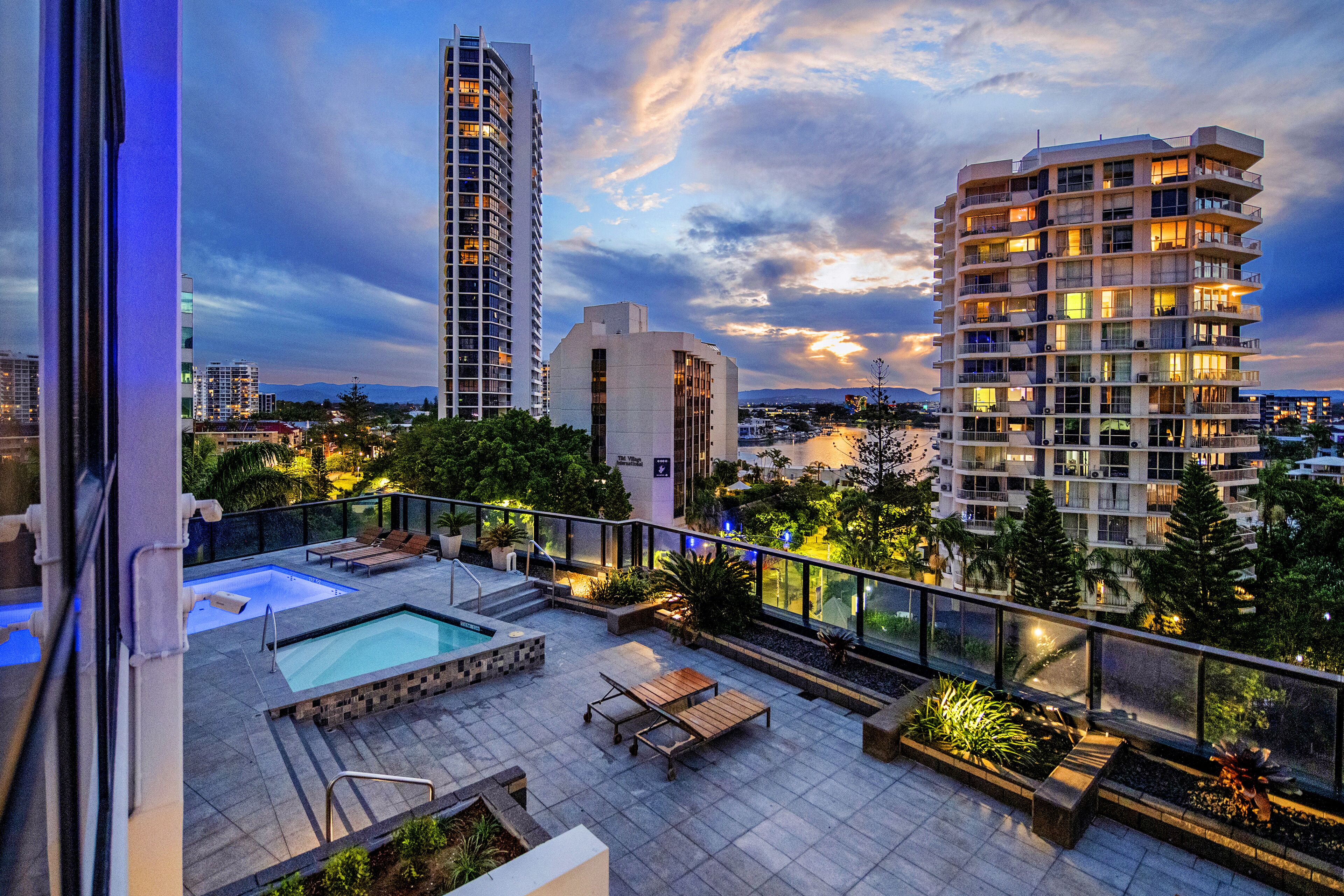 A rooftop pool area with lounge chairs and a small pool, surrounded by greenery and overlooking a cityscape with tall buildings.