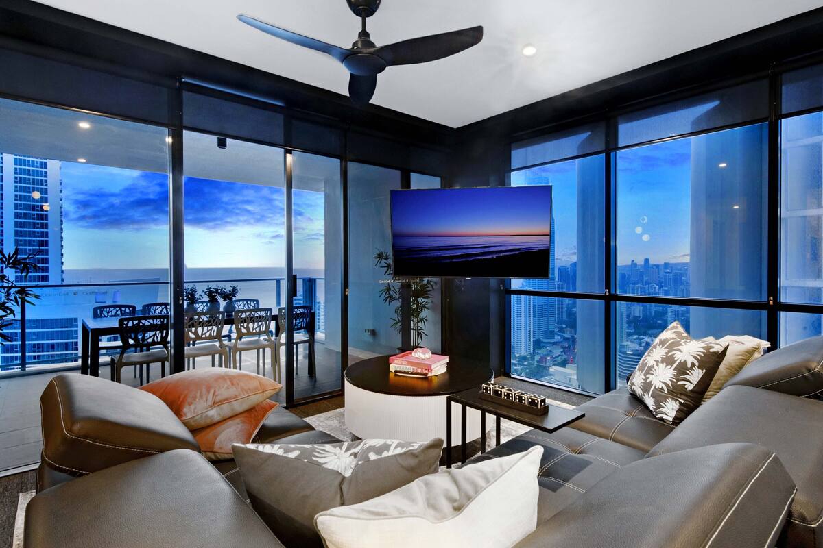 Una sala de estar moderna con un televisor grande, asientos cómodos y vistas a la ciudad a través de amplios ventanales.