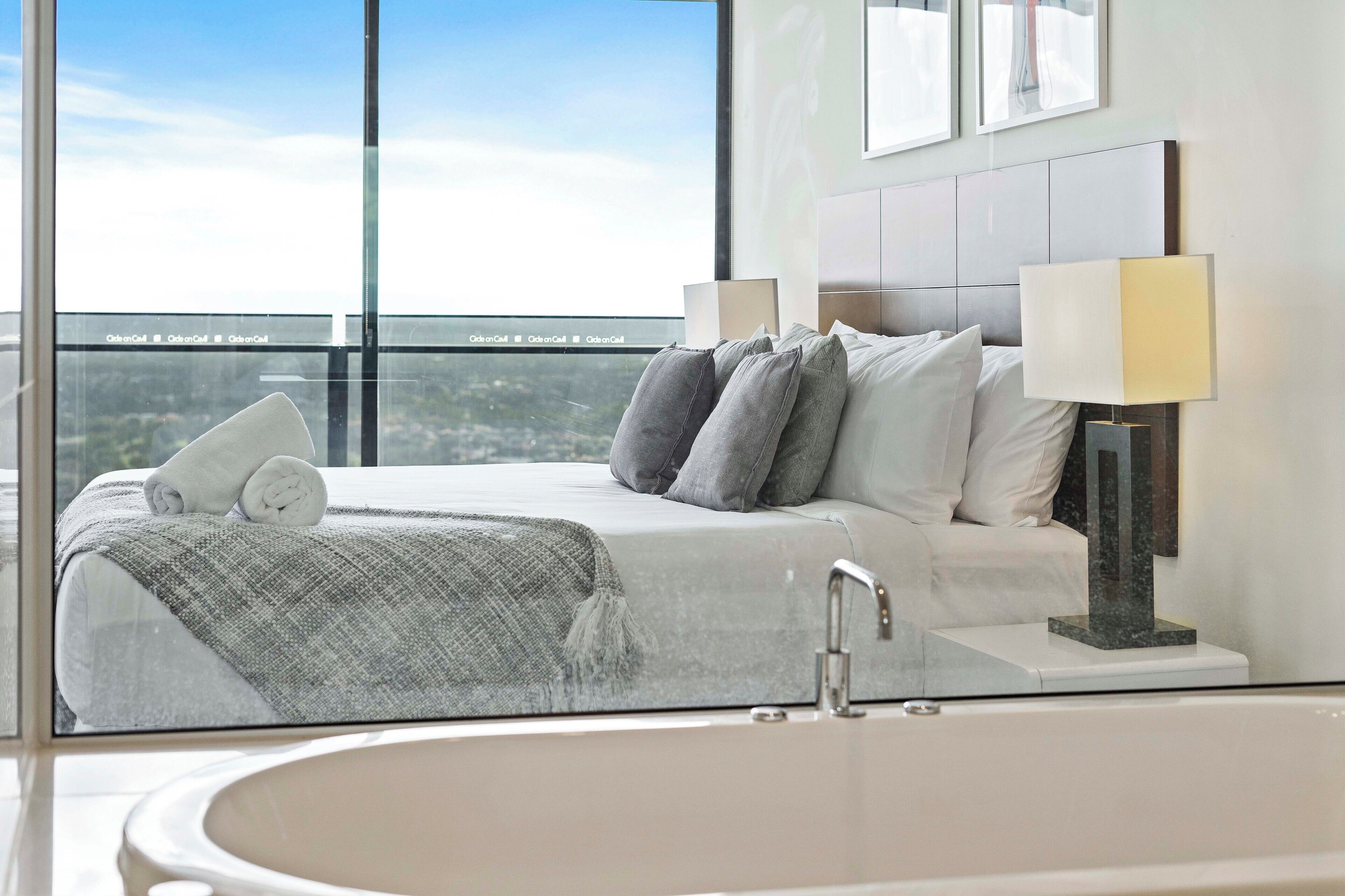 A modern bedroom with a large window, a bed with pillows, a lamp, and a bathtub.
