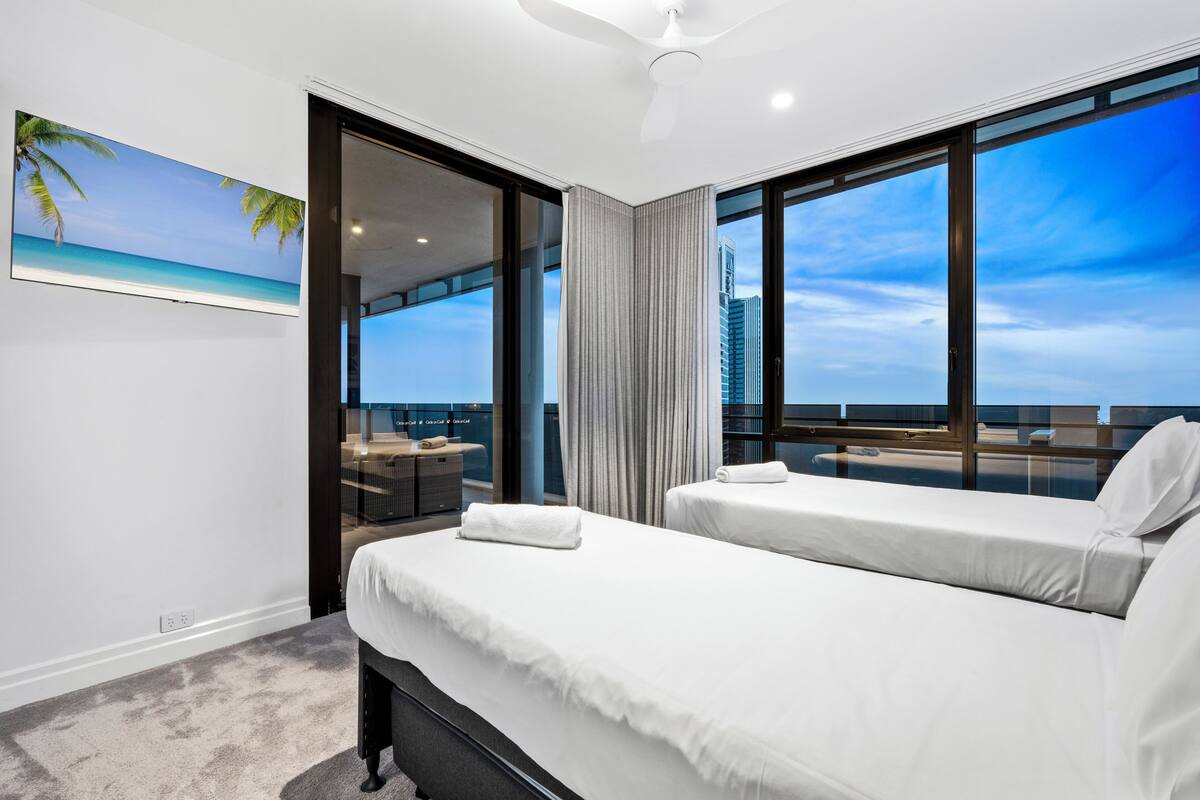 Habitación de hotel con dos camas, un ventanal con vistas, un ventilador de techo y un cuadro de una playa.