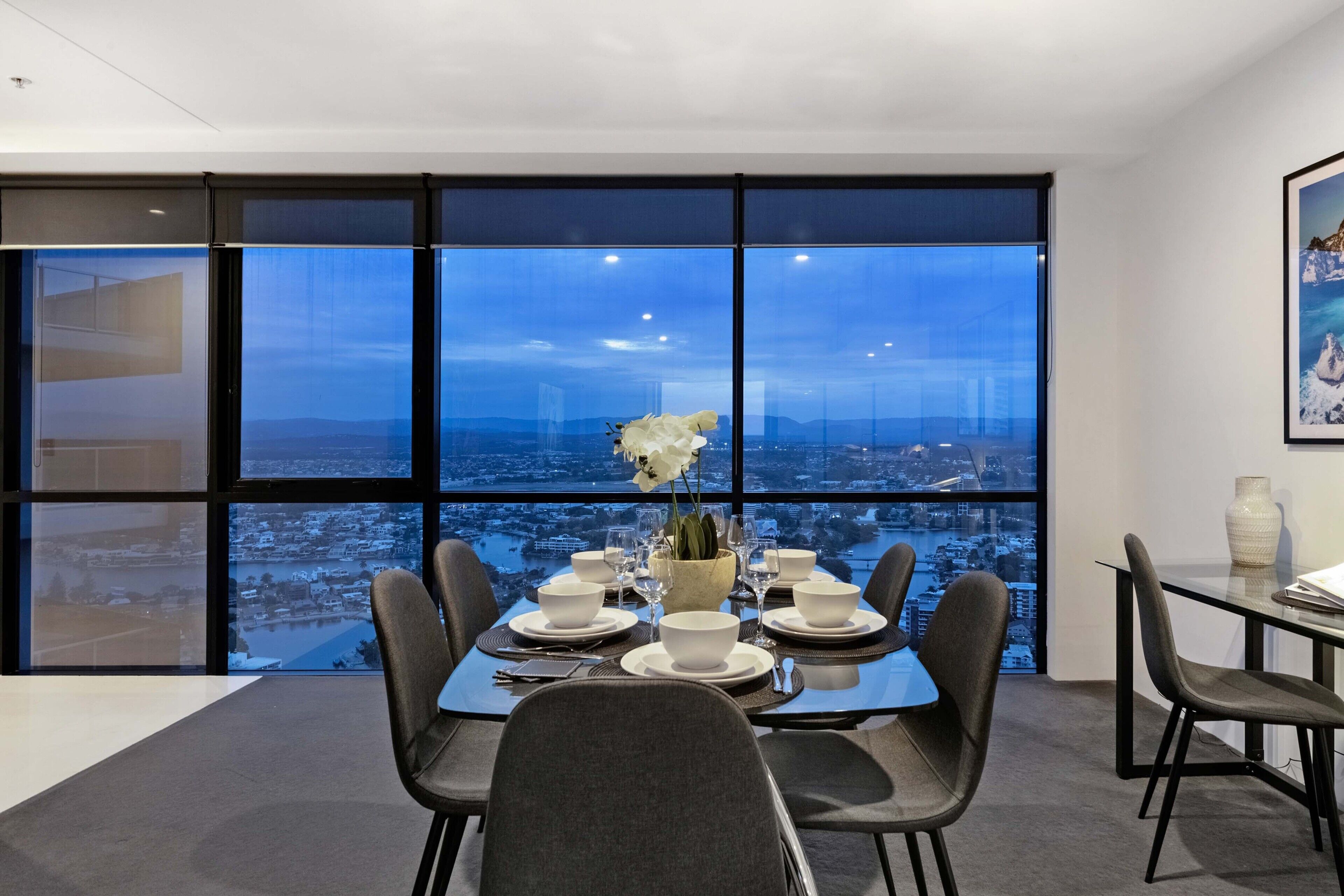 A dining area with a table set for four, large windows offering a city view, and a vase with flowers on the table.