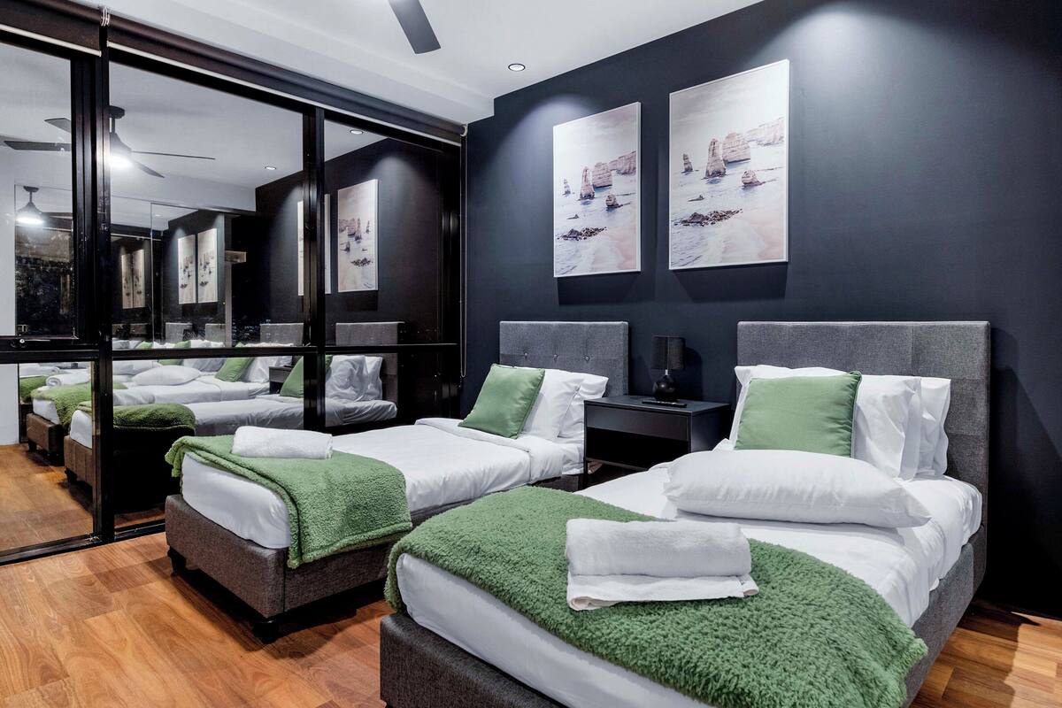 Habitación de hotel con dos camas, cada una con una manta verde, ropa de cama blanca y una mesita de noche con lámpara.