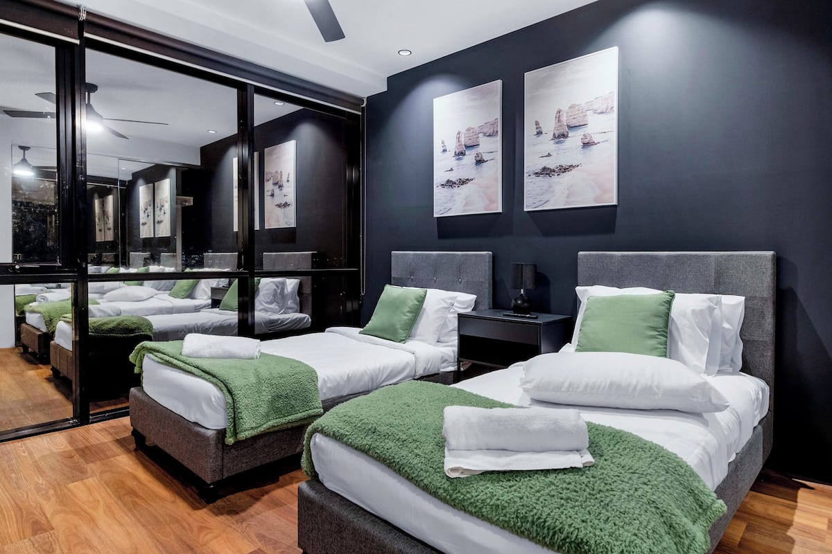Habitación de hotel con dos camas, cada una con una manta verde, ropa de cama blanca y una mesita de noche con lámpara.