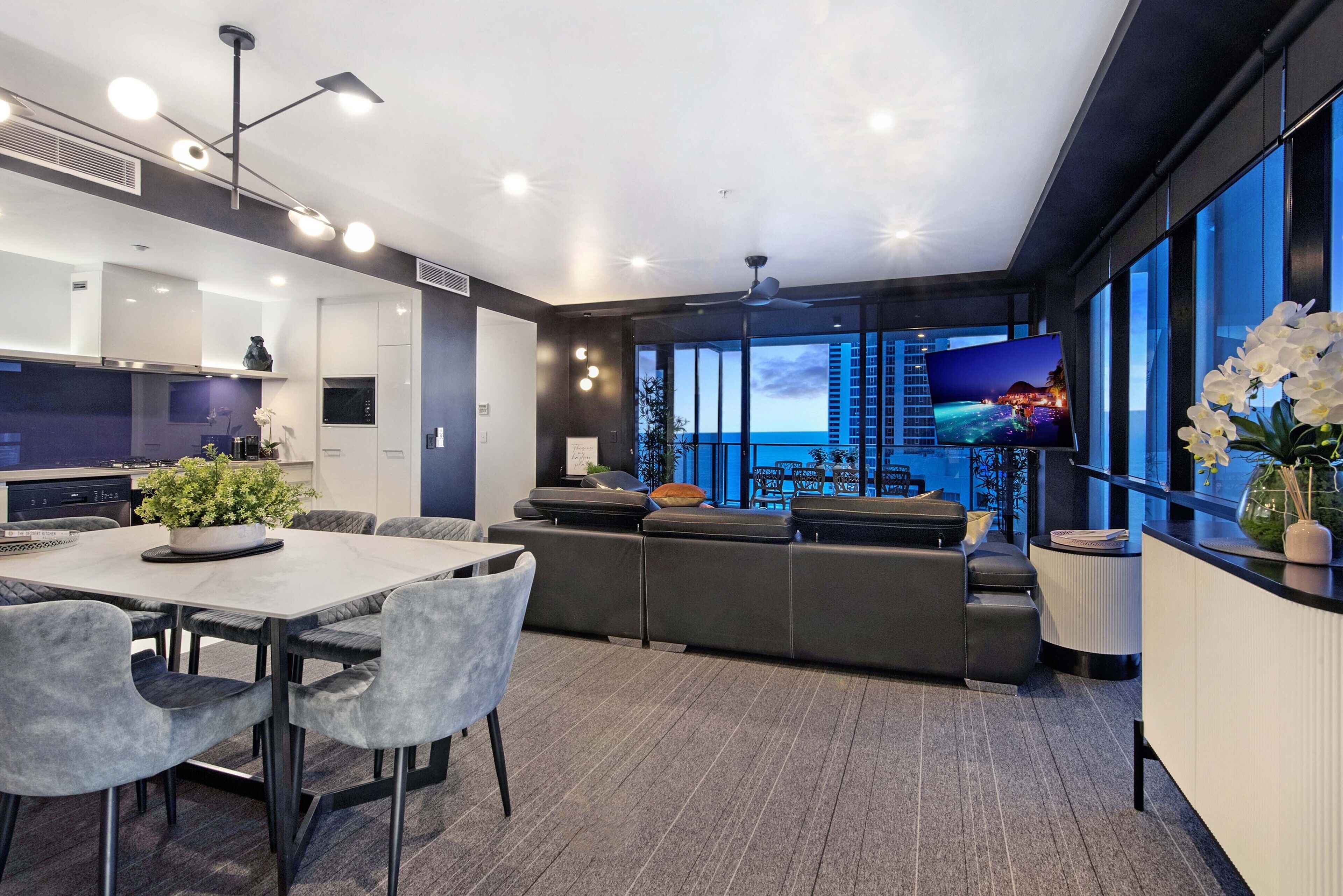 A modern kitchen with a dining area, a large flat-screen TV, and a view of the cityscape.