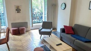 En moderne stue med en grå sofa, et træbord, en grå lænestol og et lille bord med en fjernbetjening. Der er et vindue med udsigt til træer og en balkon med gelænder af jerngitter.