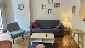 En stue med en grå sofa, et lille træ sofabord, en grå lænestol og en rund måtte. Der er billeder i rammer på væggen og et ur.