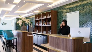 A reception area with a wooden reception desk, a vase of flowers, and a wall-mounted display case with glassware. - The Originals Boutique, Hôtel Porte de Dieppe (Saint-Aubin-sur-Scie)