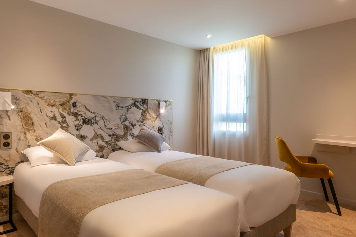 A hotel room with two beds, a marble headboard, a yellow chair, and a large window with curtains.