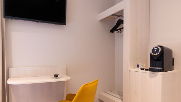 Una habitación compacta con una silla amarilla, una mesita con una taza, una cafetera y un televisor montado en la pared.