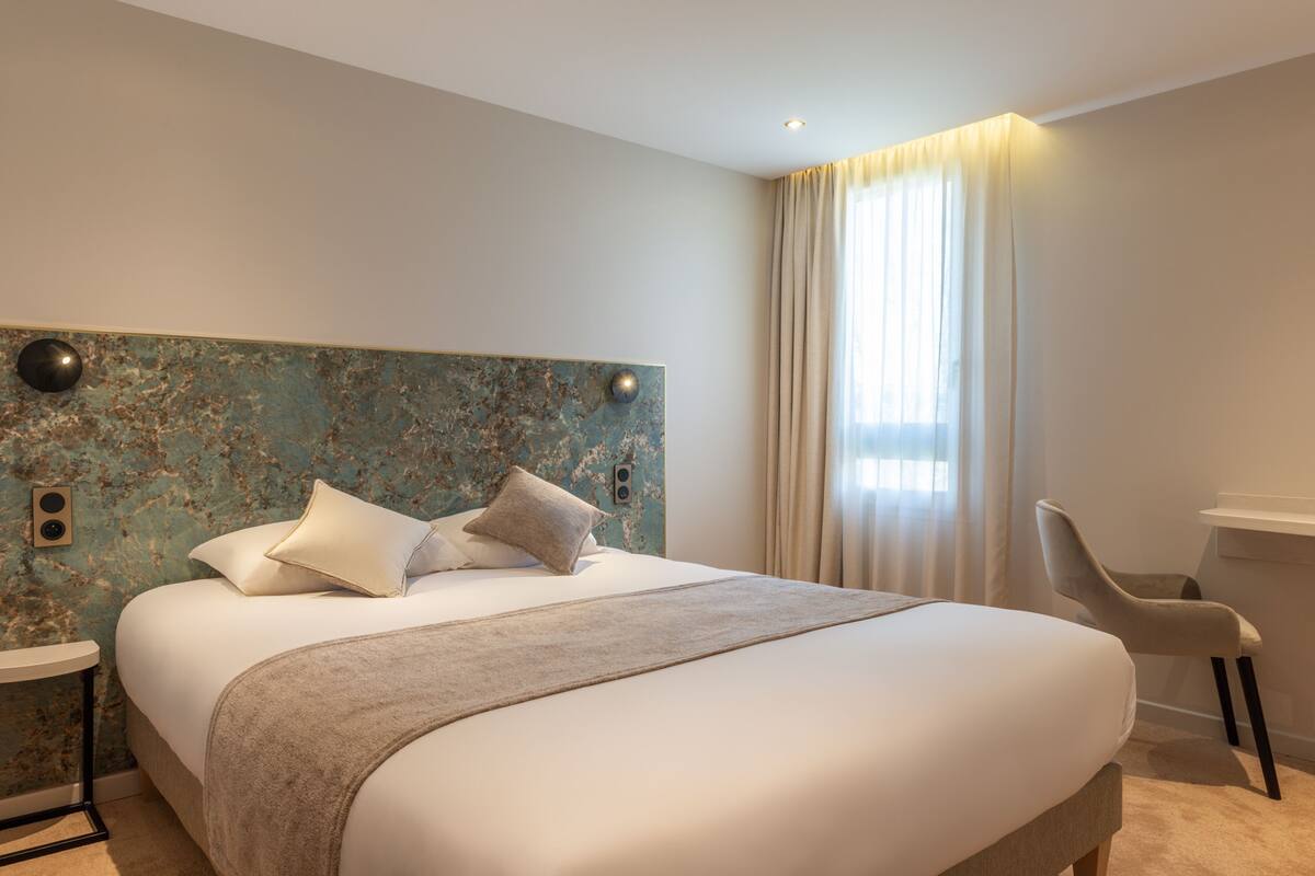 A modern hotel room with a large bed, a headboard with a textured design, a bedside table, a chair, and a window with curtains.