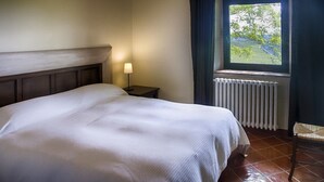 Uma cama bem arrumada em quarto com janela, luminária de cabeceira e radiador.
