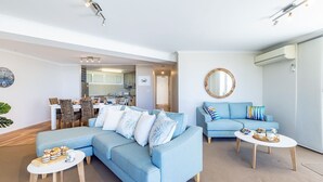 A modern living room with a sectional sofa, a blue armchair, and a coffee table. There is a kitchen in the background with a dining area. - Portofino, 2,7 Laman St - Beautifully styled unit on first floor with stunning views, Air con, Wi-Fi and close to town (Nelson Bay)