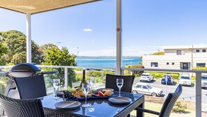 A balcony with a table set for dining, overlooking a body of water and a residential area. - Portofino, 2,7 Laman St - Beautifully styled unit on first floor with stunning views, Air con, Wi-Fi and close to town (Nelson Bay)
