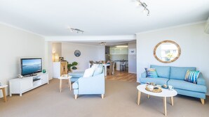 A modern living room with a TV, sofa, coffee table, and dining area. - Portofino, 2,7 Laman St - Beautifully styled unit on first floor with stunning views, Air con, Wi-Fi and close to town (Nelson Bay)