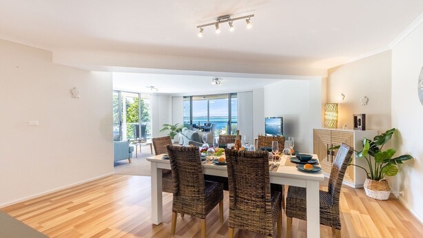 A dining area with wicker chairs, a white table, and a view of the sea through large windows. - Portofino, 2,7 Laman St - Beautifully styled unit on first floor with stunning views, Air con, Wi-Fi and close to town (Nelson Bay)