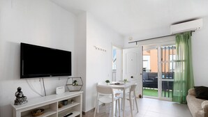 Un salon équipé d’un téléviseur à écran plat, d’une table à manger blanche avec des chaises, d’un canapé et d’une porte-fenêtre coulissante donnant sur un balcon.