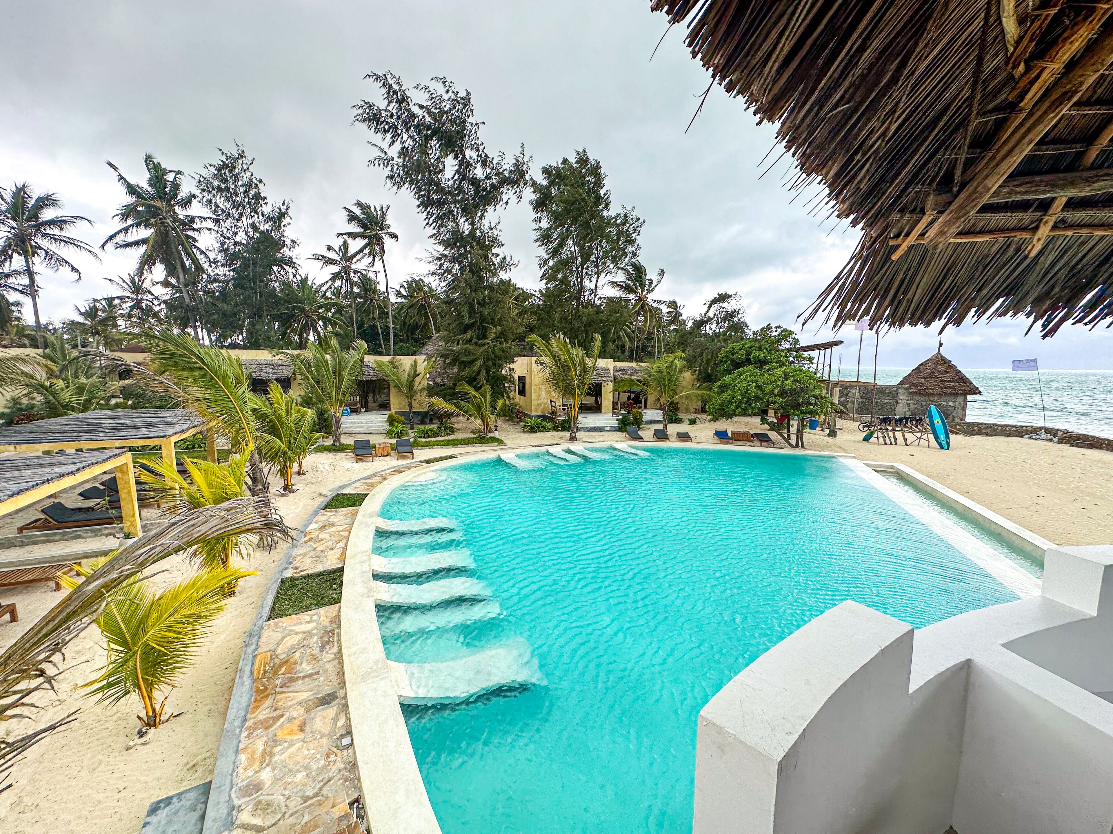 A pool area with a curved edge, surrounded by lounge chairs and a thatched-roof structure.