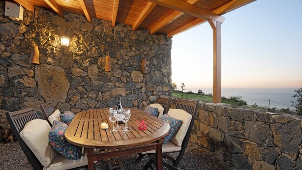 Un muro in pietra con una pergola in legno e un tavolo rotondo in legno con sedie.