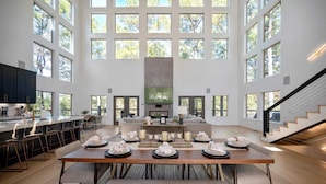 A spacious dining area with a long table set for a meal, a kitchen with modern appliances, and a large windowed wall providing natural light.