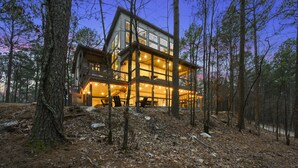 A large, multi-story wooden cabin with a balcony, surrounded by trees and a forest floor covered with leaves and pine needles.