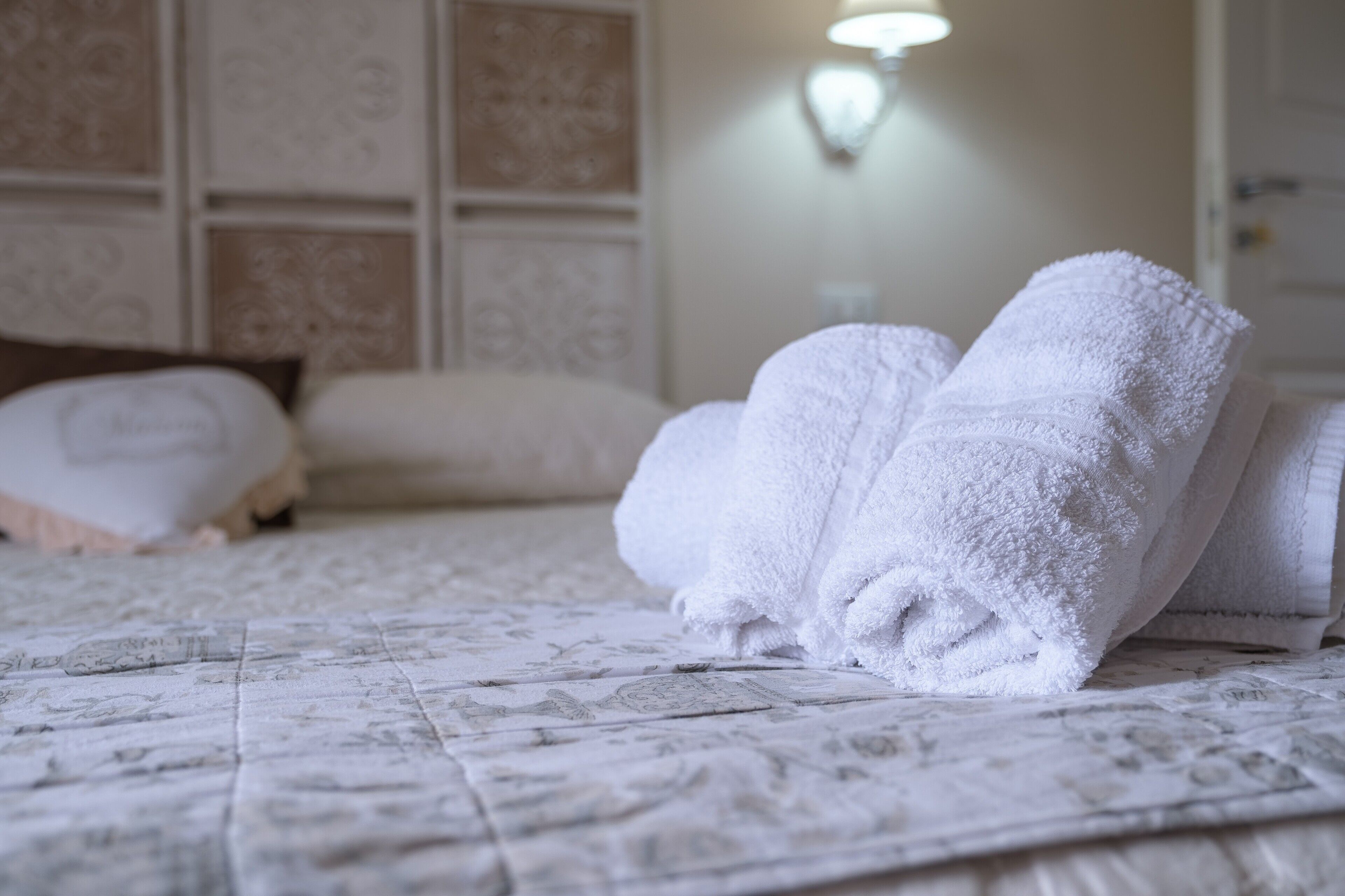 Un lit bien fait, avec des serviettes blanches pliées en rouleau, une couvre-lit à motifs et un luminaire fixé au mur.
