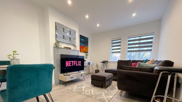 En moderne stue med et fladskærms TV, der viser 'NETFLIX', en teal farvet lænestol, en brun sofa og et spiseområde med bord og stole.
