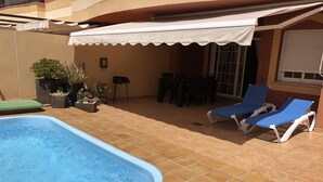 Una piscina con piso de baldosas, un porche cubierto con sillas y mesas, y un toldo blanco.