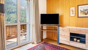 A room with a large window, a flat-screen TV, a wooden cabinet, and a red patterned rug. - Holiday Apartment "Salman" with Mountain View, Balcony & Wi-Fi (Ortisei)