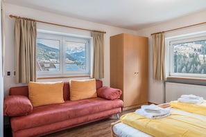 A room with a red sofa, a bed with yellow bedding, a wooden wardrobe, and a view of mountains through the windows.