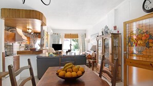 A dining area with a wooden table, chairs, a bowl of fruit, and a kitchen in the background.
