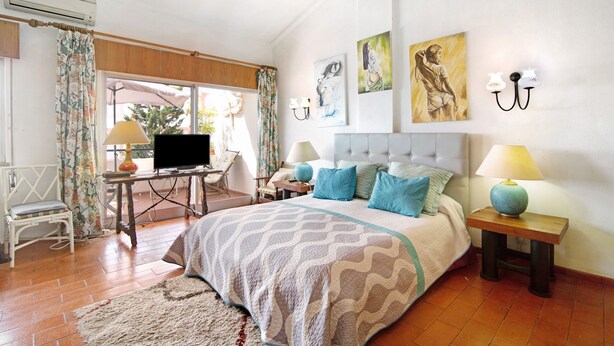 A bedroom with a bed, two bedside tables with lamps, a chair, a desk with a TV, and artwork on the walls.