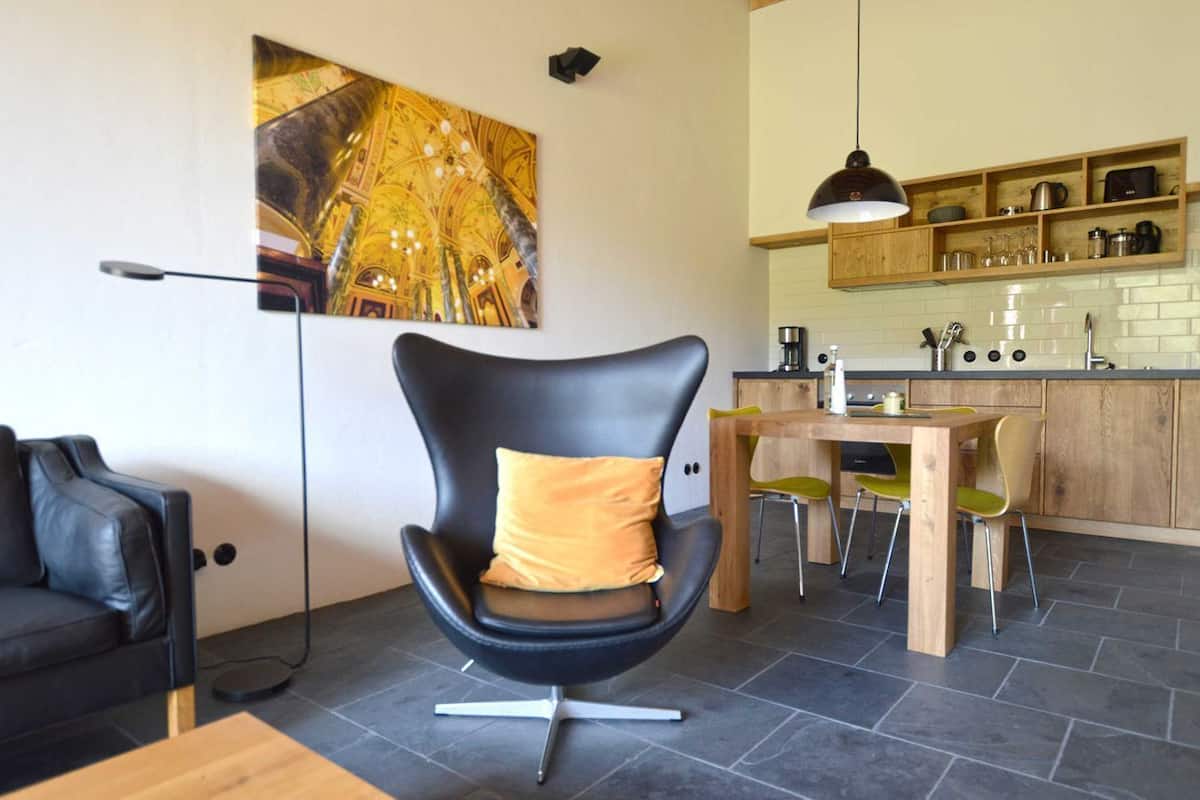 Un salon moderne avec un fauteuil en cuir noir, une table à manger en bois et un coin cuisine doté de meubles en bois et d’un luminaire suspendu.